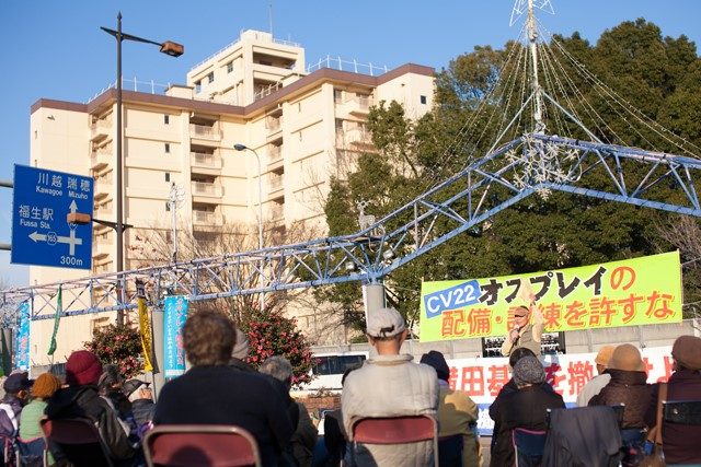 ▲12月18日に開催された「横田基地の撤去を求める座り込み行動」には、主催者発表によれば106名が参加した。集会が行われたフレンドシップパーク（東京・福生市）の車道を一本挟んだ塀の向こう側に、横田基地が広がる。写真の背景に写るビルと樹木は基地の敷地内のもの。